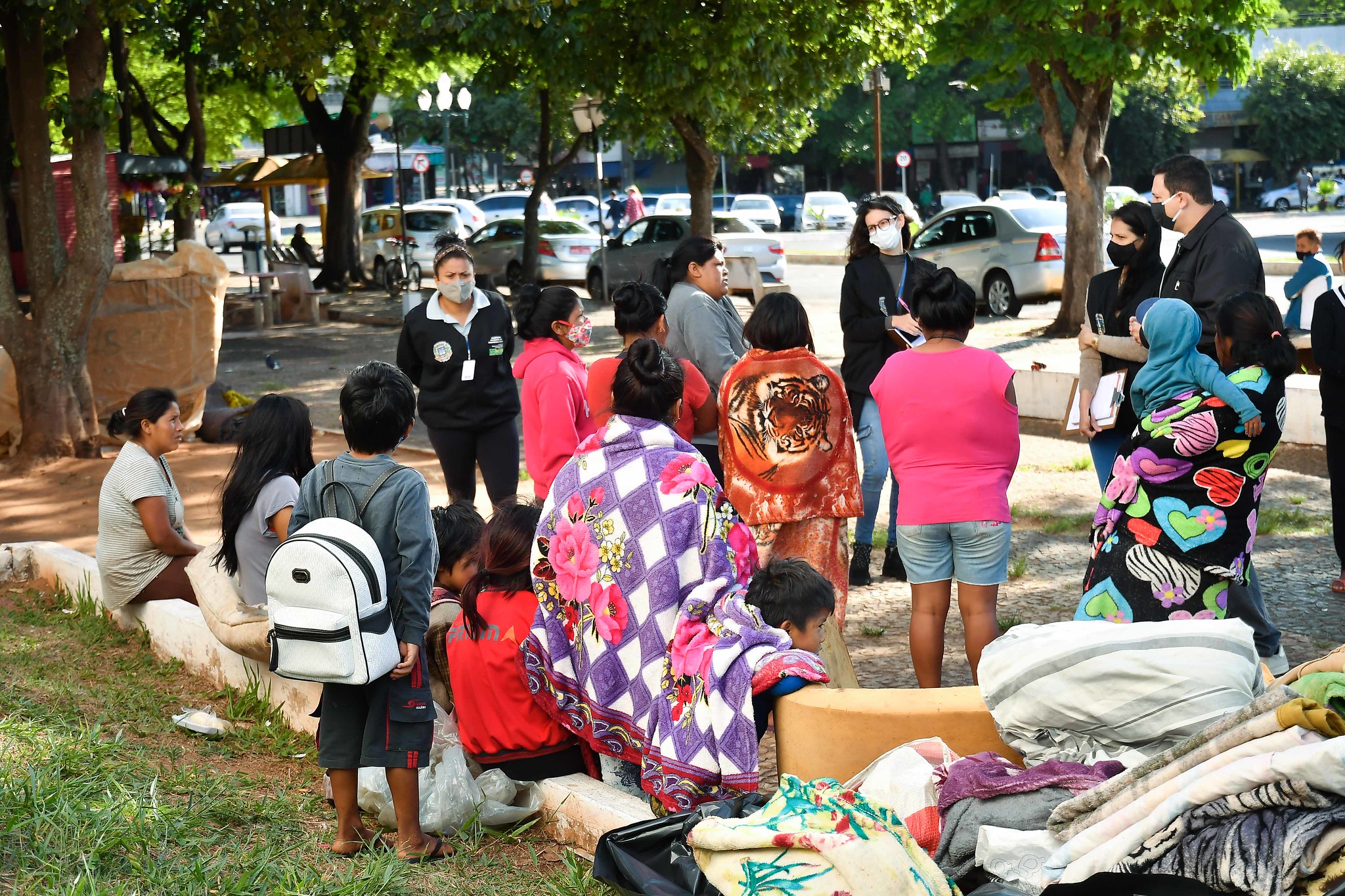Várias pessoas envoltas em cobertores e roupas de frio paradas em volta de uma praça, sendo abordadas por homens e mulheres da assistência social