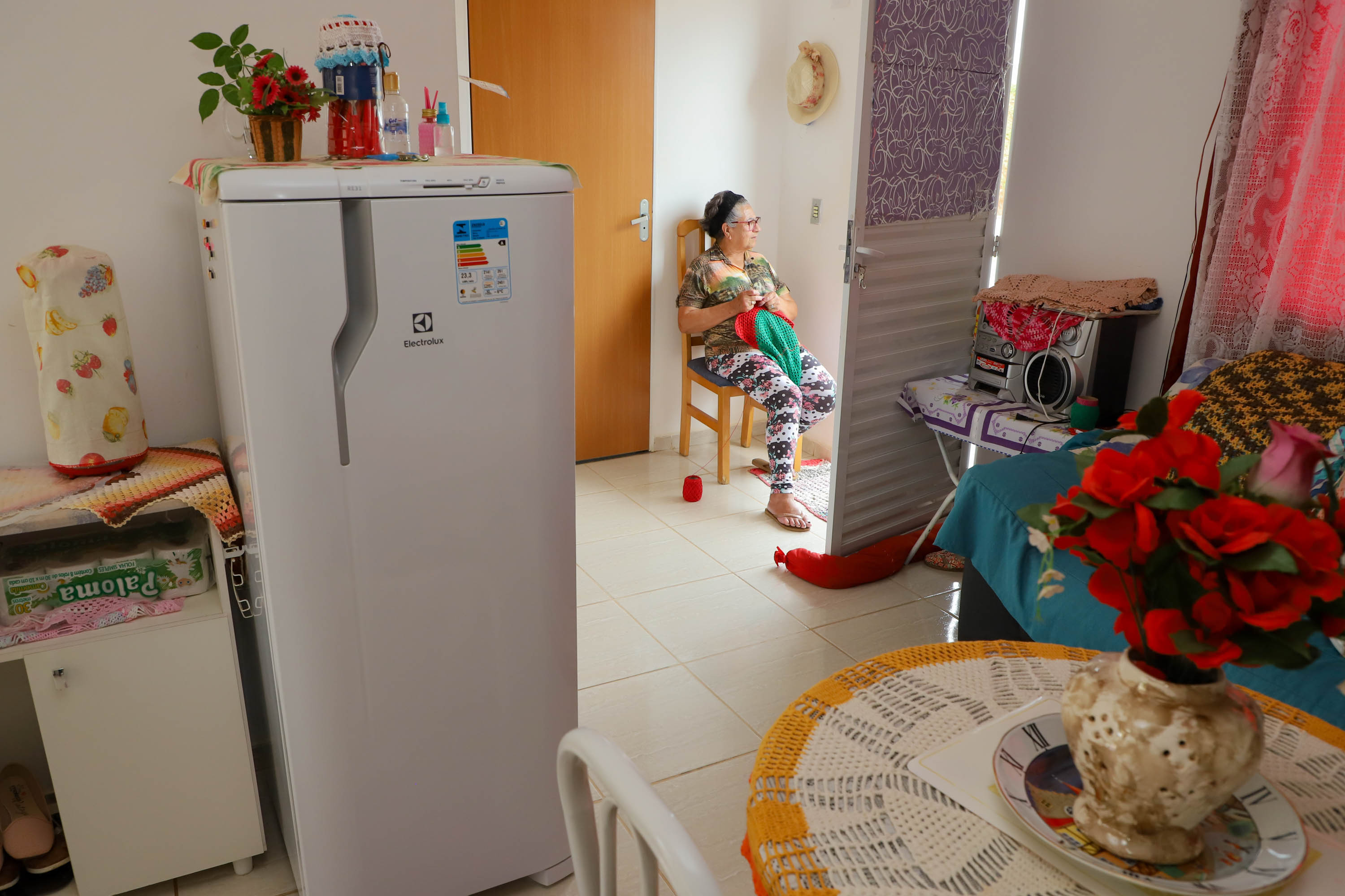 Imagem de uma senhora sentada na cadeira à porta de sua casa, mobiliada com geladeira, mesa, aparelho de som e flores