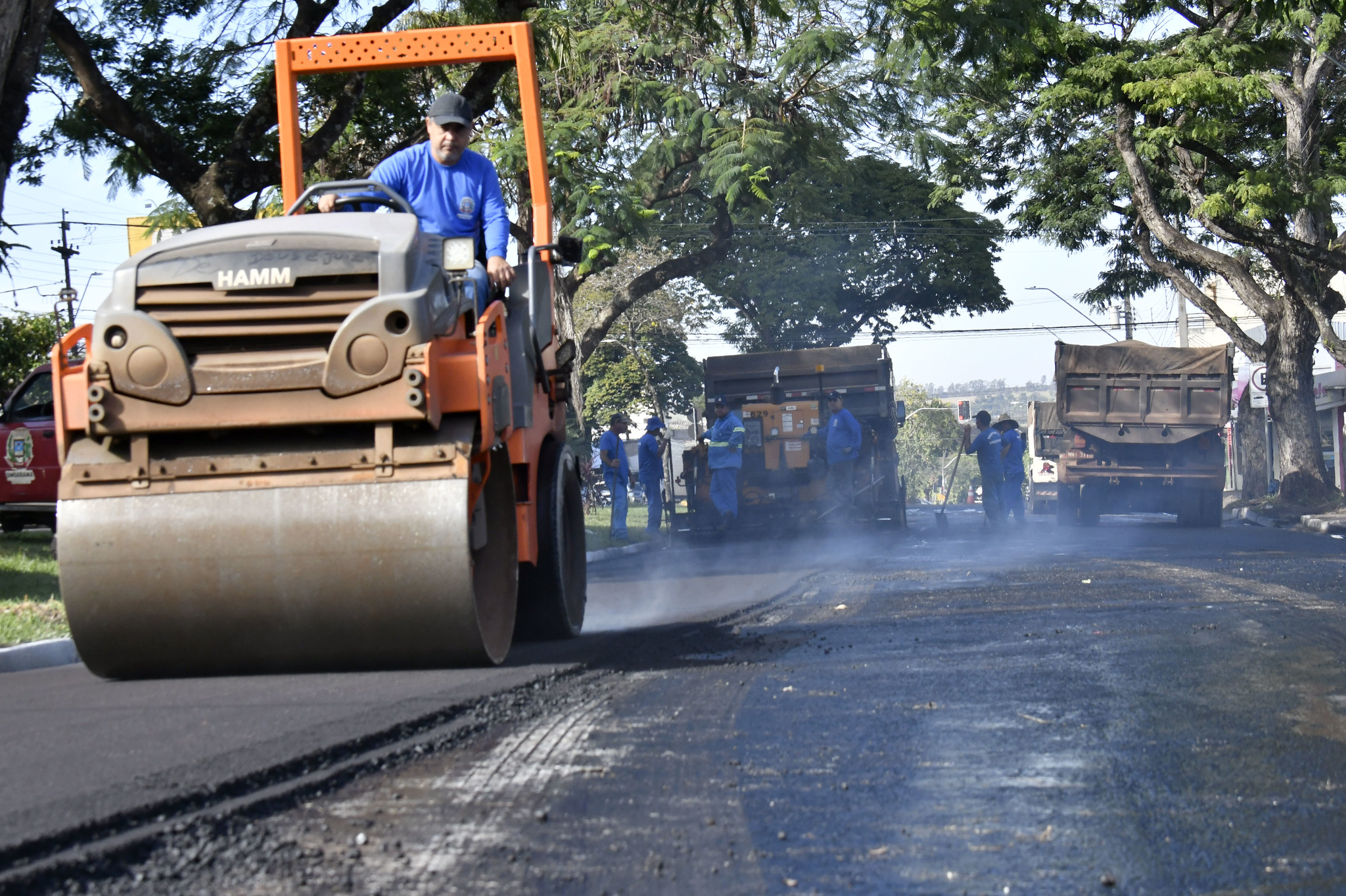 Veículo rolo compressor compacta capa de recape asfáltico aplicada sobre asfalto deteriorado; ao fundo, homens trabalhando com uniformes azuis e um caminhão parado