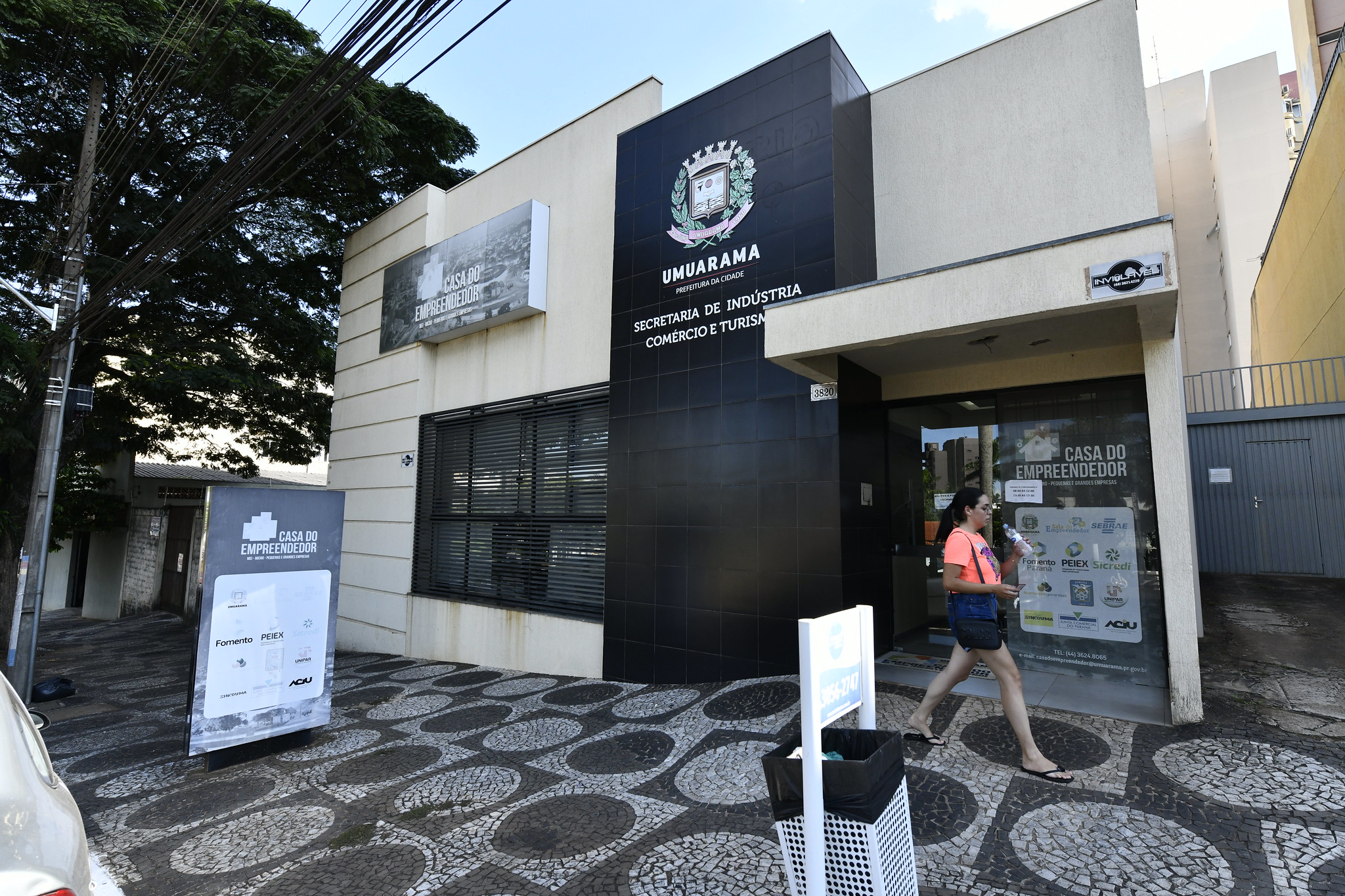 Imagem da fachada da Casa do Empreendedor, um prédio em alvenaria com portas e janelas em vidro, uma calçada em petit pave e uma mulher caminhando na frente