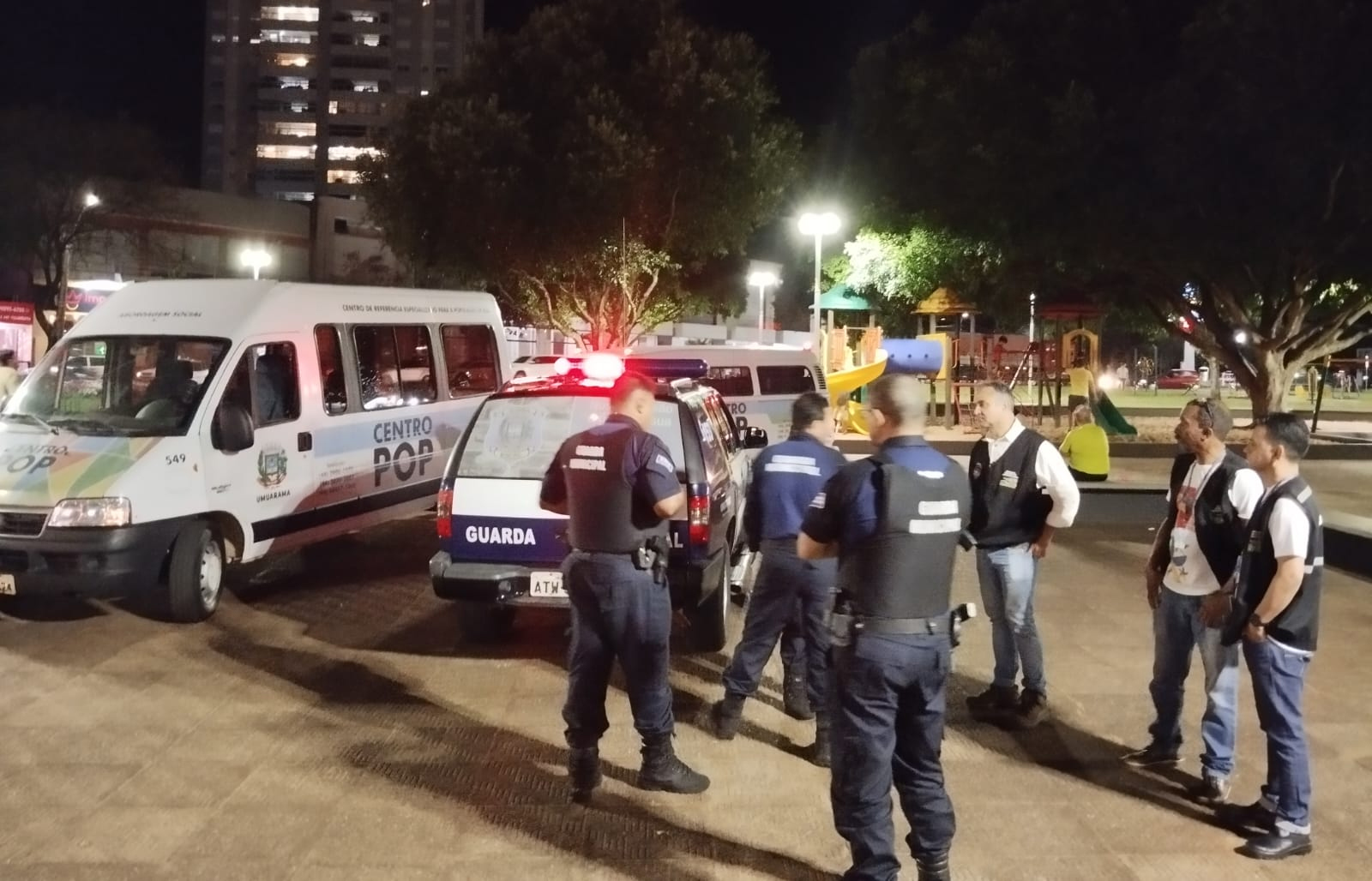 Homens, agentes da guarda municipal uniformizados e com coletes reunidos em uma praça, ao lado de veículos e viaturas policiais