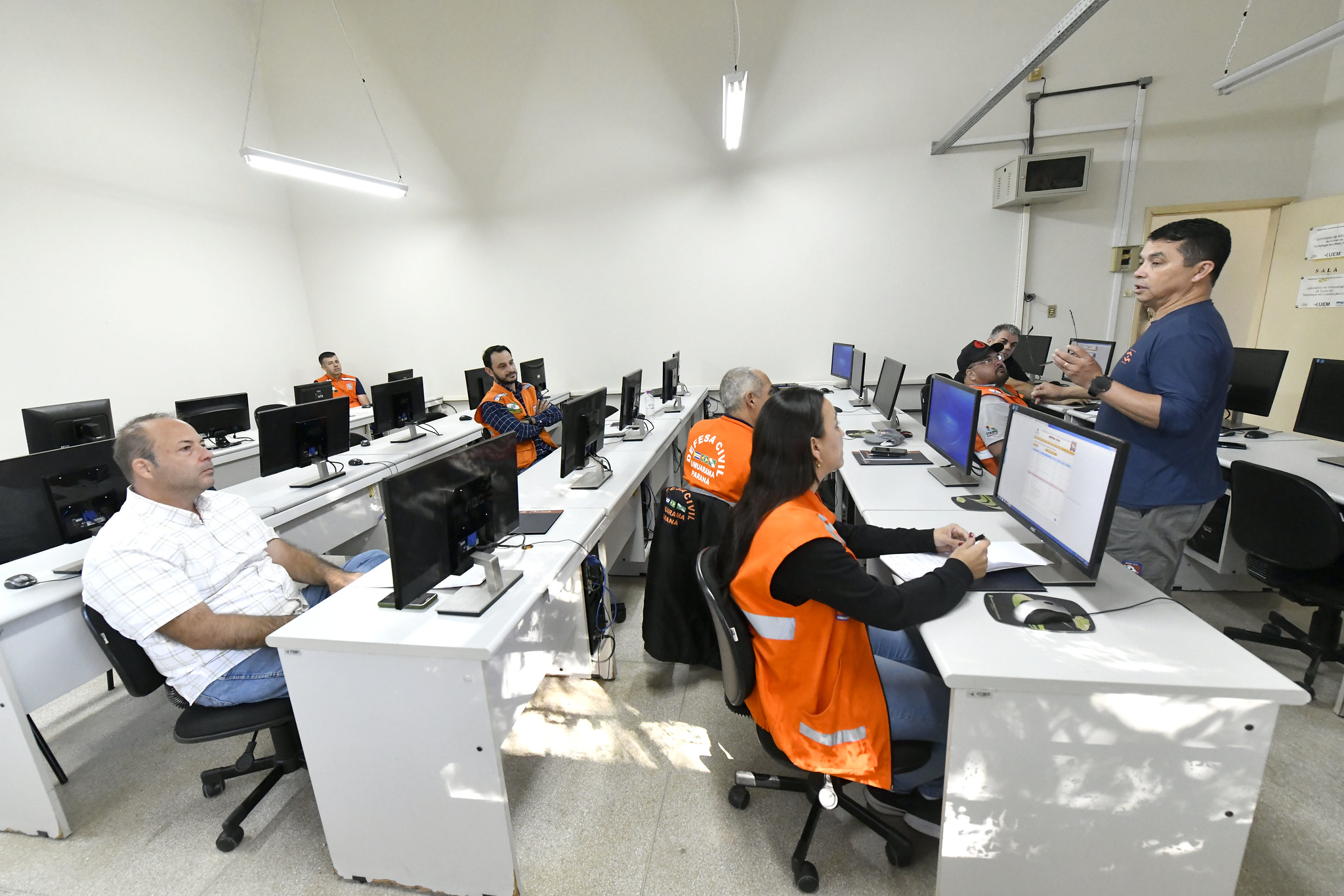 Curso com agentes das defesa civil usando jaquetas de cor laranja, reunidos em um laboratório de informática, à frente do instrutor em pé da parte da frente da sala