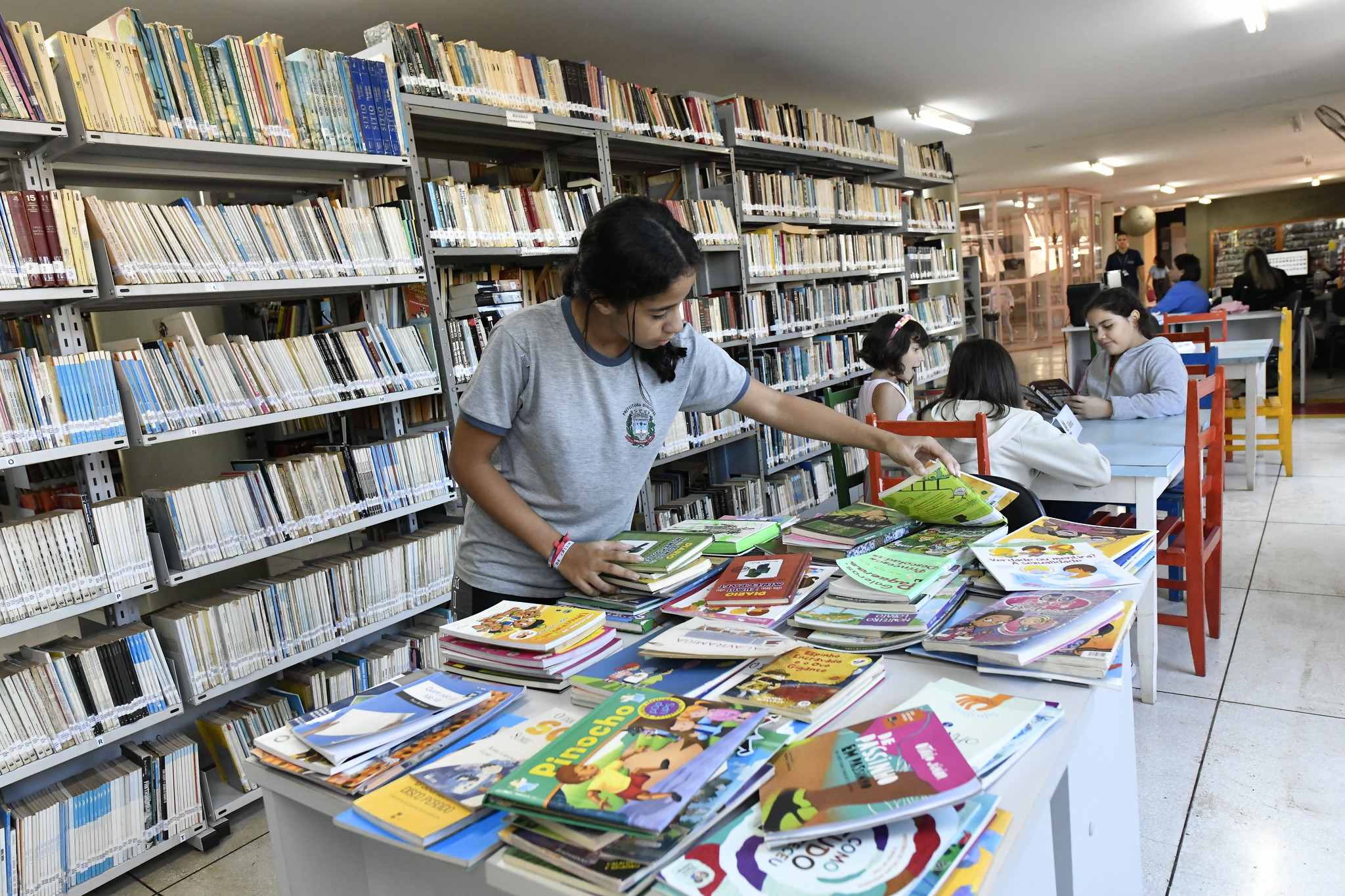 alunos manuseiam livros na biblioteca municipal, diante de estantes de livros