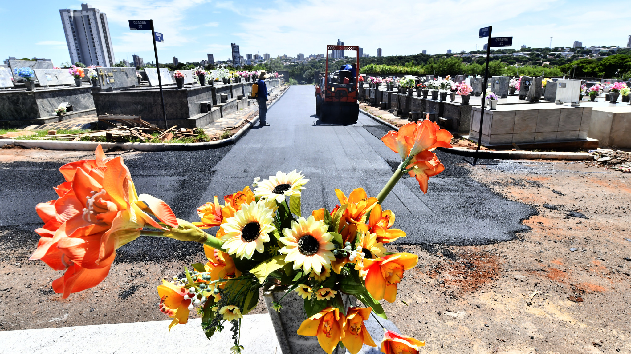 vaso de flores coloridas em primeiro plano; ao fundo, máquinas da prefeitura realizam a pavimentação de acesso no cemitério