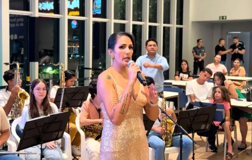organizadora do evento fala ao microfone, diante da orquestra (ao fundo) em ação no shopping