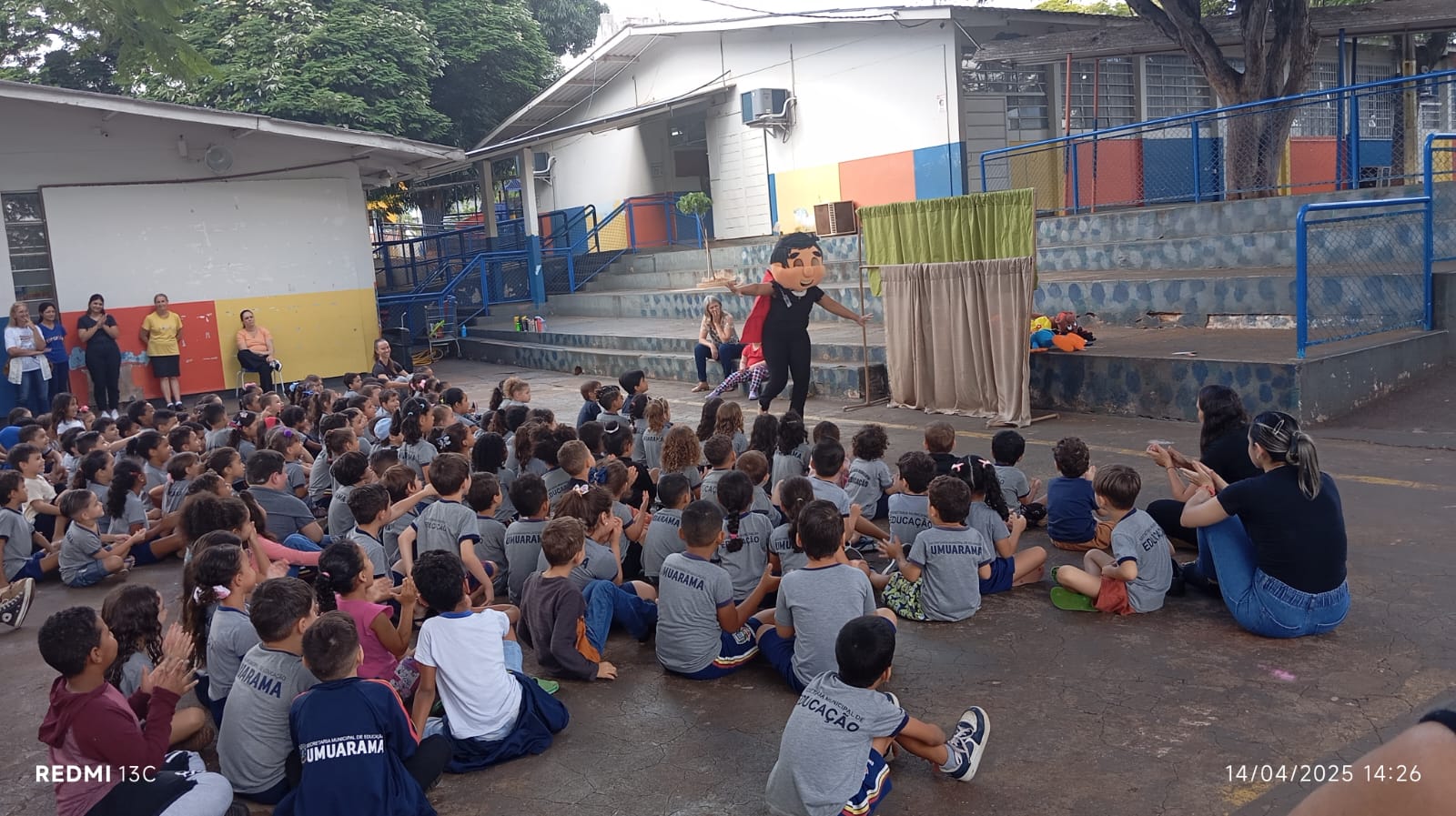 crianças prestigiam teatro sobre a dengue sentadas no pátio da escola