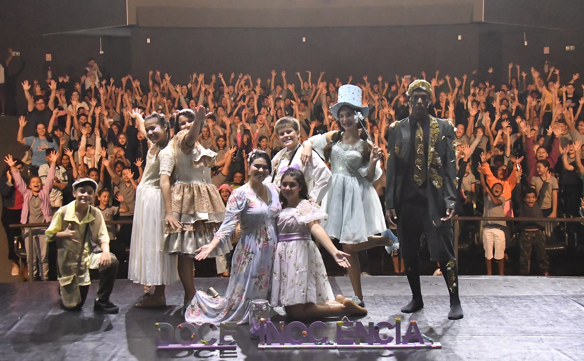 artistas no palco e plateia ao fundo, lotando cadeiras do teatro