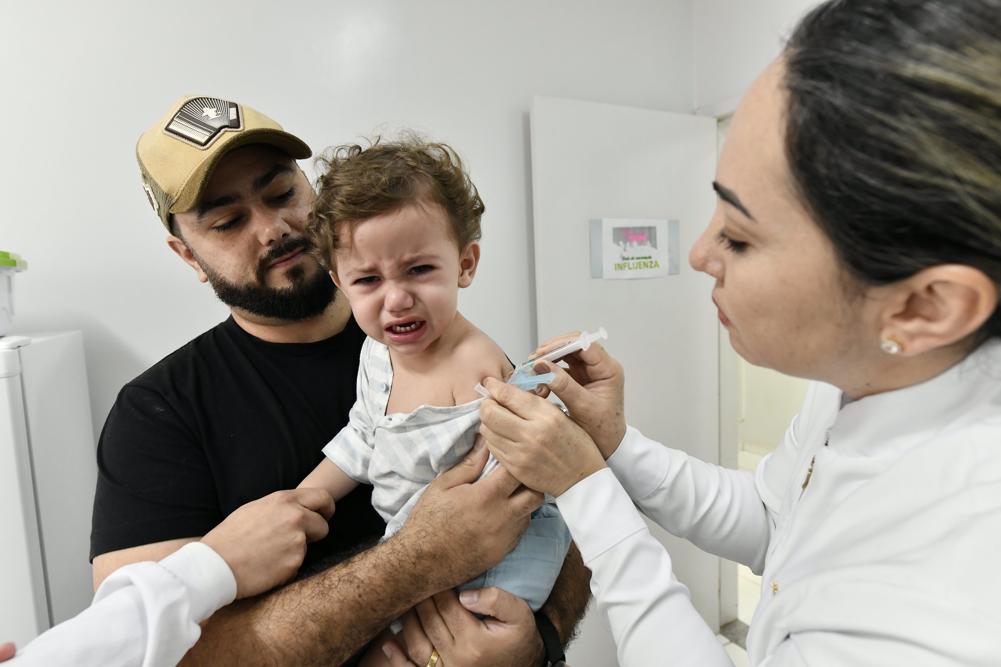 criança chora ao receber vacina no braço, amparada pelo pai