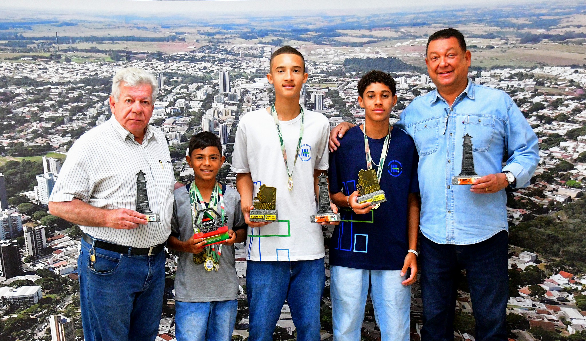 prefeito e secretário de esportes posam para foto com atletas jovens exibindo troféus e medalhas