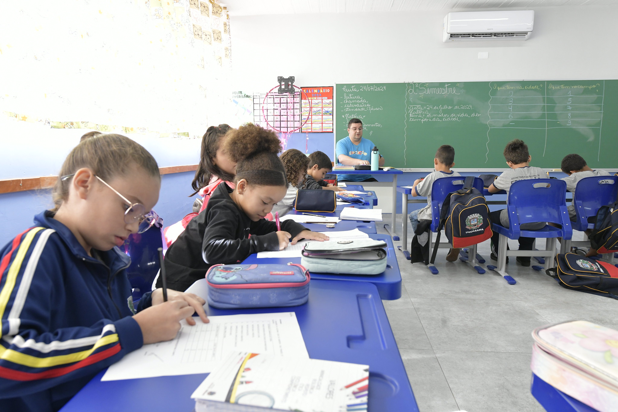 sala de aula com crianças estudando e professor ao fundo