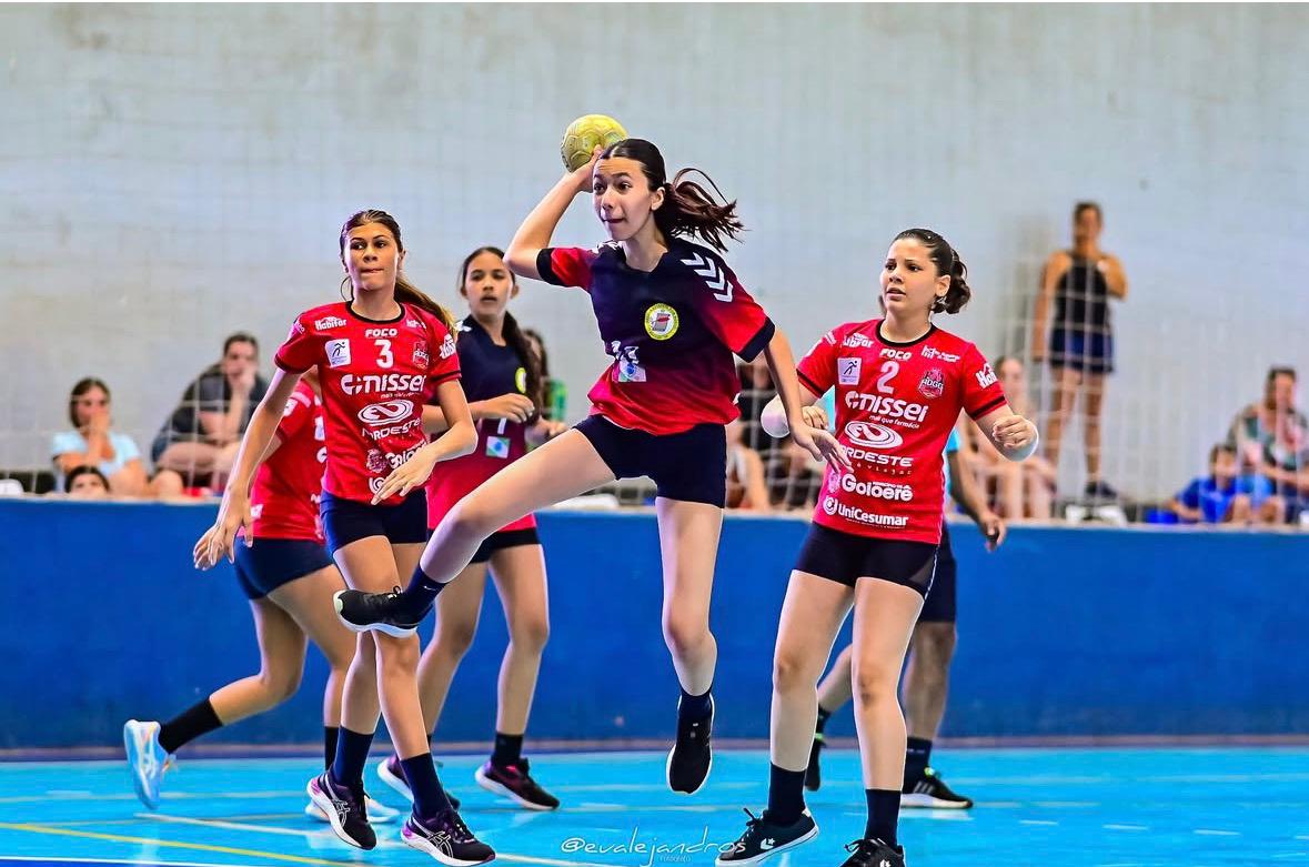 atleta feminina salta para arremessar a bola de handebol diante das adversárias