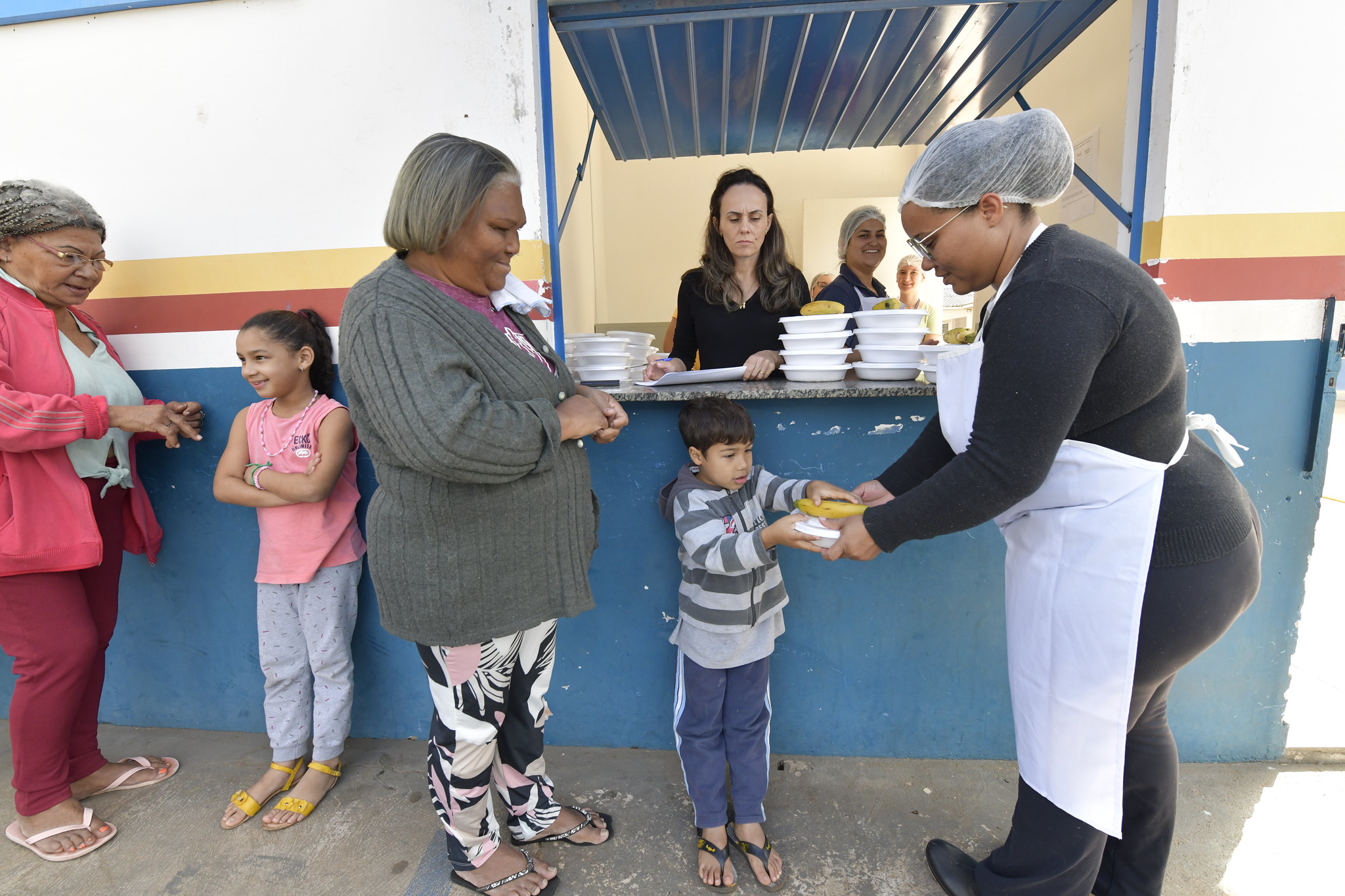 merendeira entrega refeição em marmita para criança em fila de espera