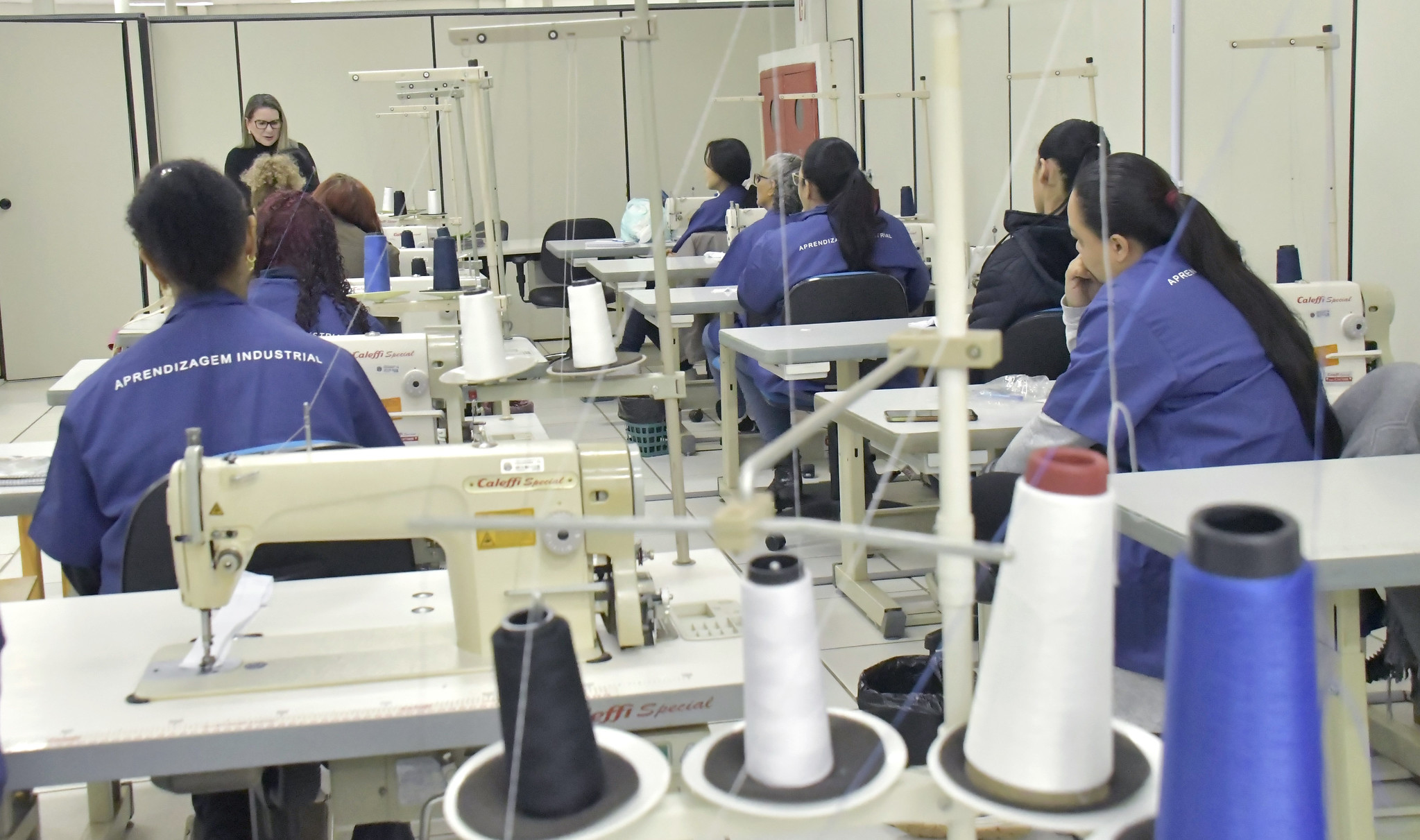 professora dá aula de costura para alunas em ambiente com máquinas industriais