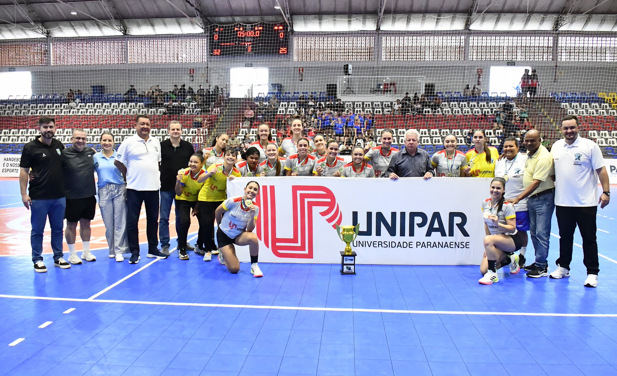 equipe comemora o título com faixa do patrocinador no ginásio de esportes