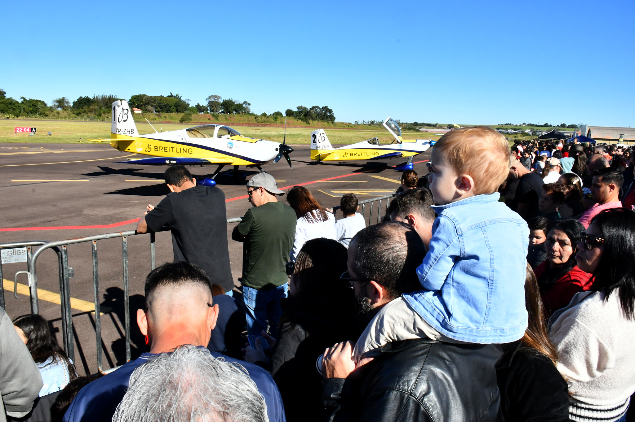 criança no colo do pai e grande público observam aviões na pista do aeroporto