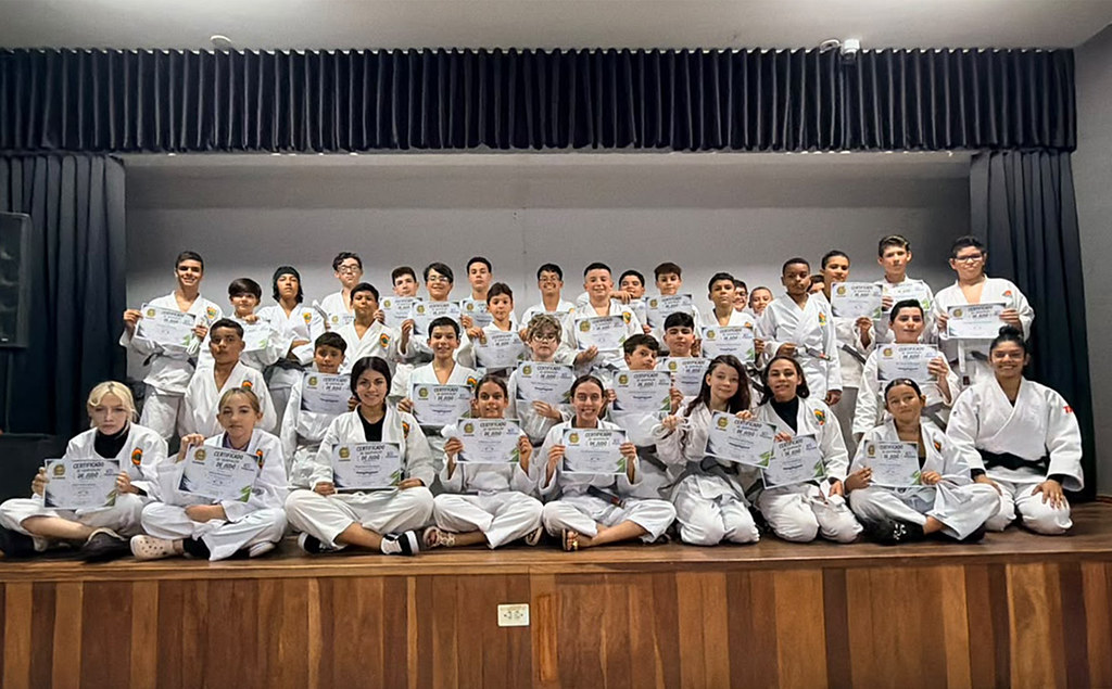 Cinco jovens judocas recebendo certificado e uma delas com um familiar ao lado durante formatura da Oficina de Judô no CEJU