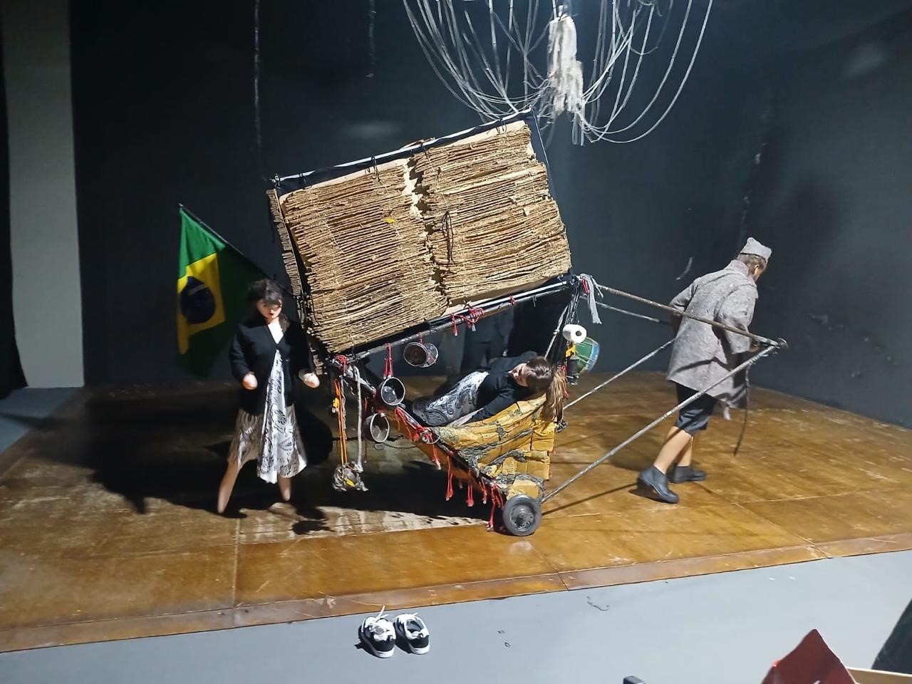 Encenação peça teatral BR Felicidade, que será apresentada no Centro Cultural Vera Schubert, em Umuarama, neste sábado (25). Na cena, um homem puxando carrinho carregado com papelão e mulher atrás com bandeira do Brasil