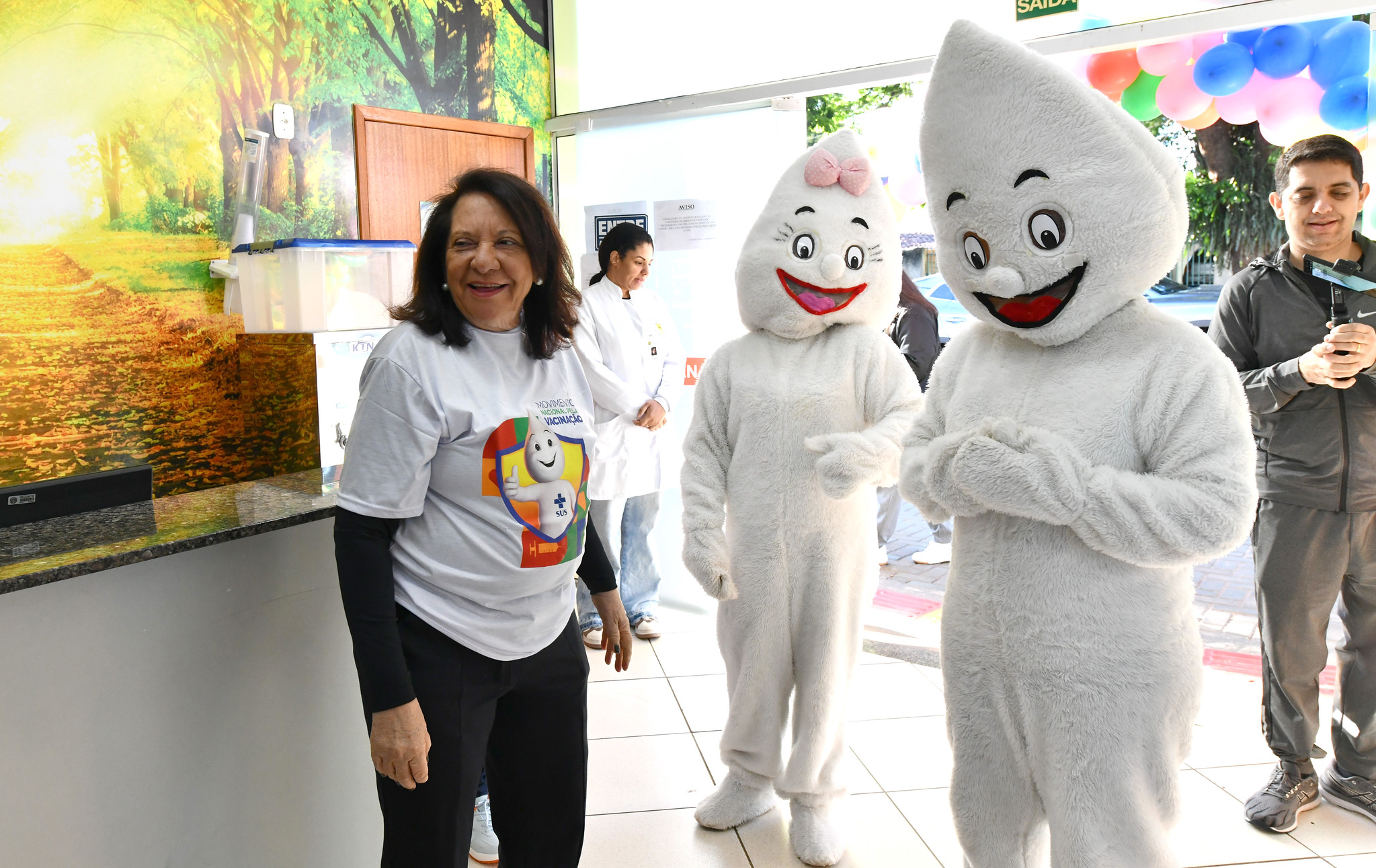 secretária da saúde visita posto de vacinação, ao lado dos personagens Zé e Maria Gotinha